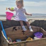 A toddler in a white dress sits on a wooden beach toy library box on Swansea promenade holding a spade, with buckets and spades inside the box and the beach behind