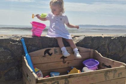 A toddler in a white dress sits on a wooden beach toy library box on Swansea promenade holding a spade, with buckets and spades inside the box and the beach behind