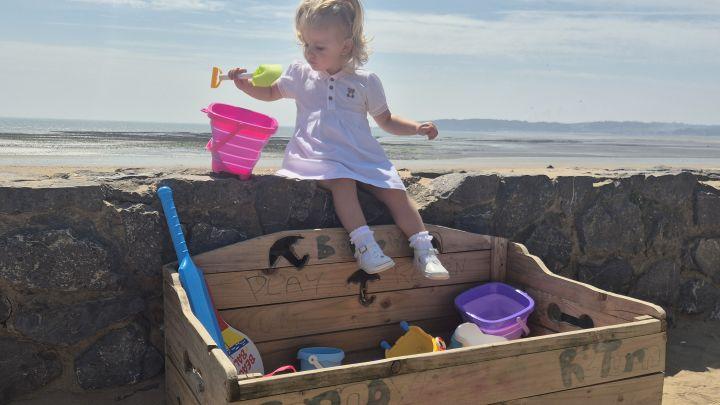 A toddler in a white dress sits on a wooden beach toy library box on Swansea promenade holding a spade, with buckets and spades inside the box and the beach behind