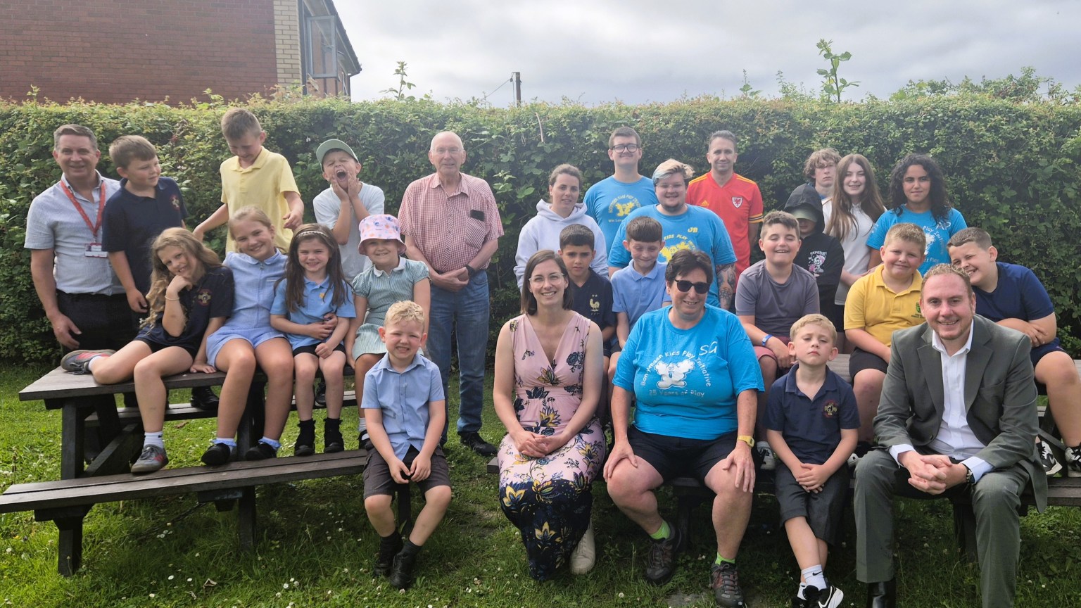 A large group of children, volunteers and councillors sitting on picnic benches in front of a hedge at the Bonymaen Kids Play Initiative