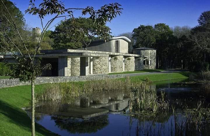 Coychurch Crematorium's low stone and concrete buildings reflected in a pond fringed with reeds, surrounded by trees under a blue sky