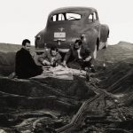 A black and white collage of three men picnicking beside a vintage car on a barren hilly landscape crossed by oil pipelines.