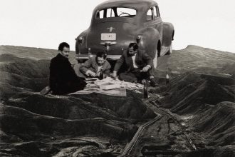 A black and white collage of three men picnicking beside a vintage car on a barren hilly landscape crossed by oil pipelines.