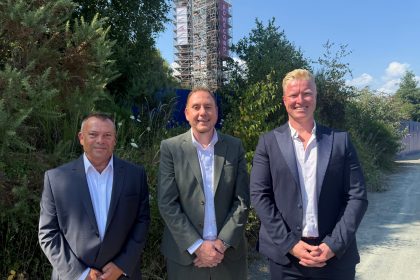 David James, Rob Stewart and Ross Miller standing in front of greenery at the Hafod-Morfa Copperworks, with the scaffolded engine house tower behind them