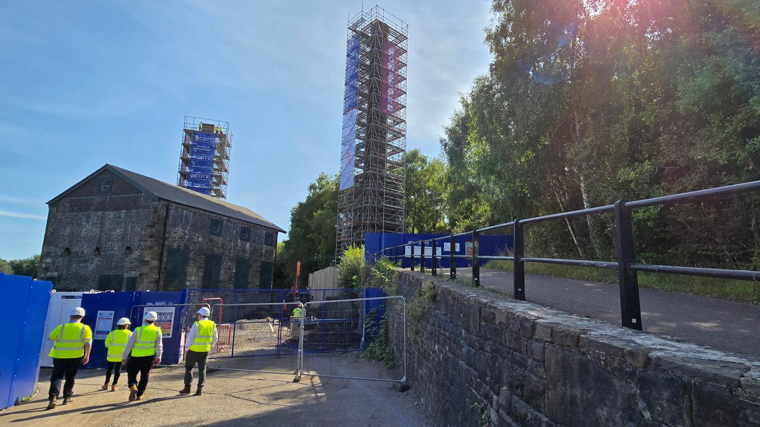 The two scaffolded chimneys at the Hafod-Morfa Copperworks with the stone engine house in the foreground and workers in hi-vis walking through the site