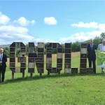 A group of people beside large wooden letters spelling GNOLL filled with bug hotels, holding a bilingual Green Flag Award 2026/27 banner.