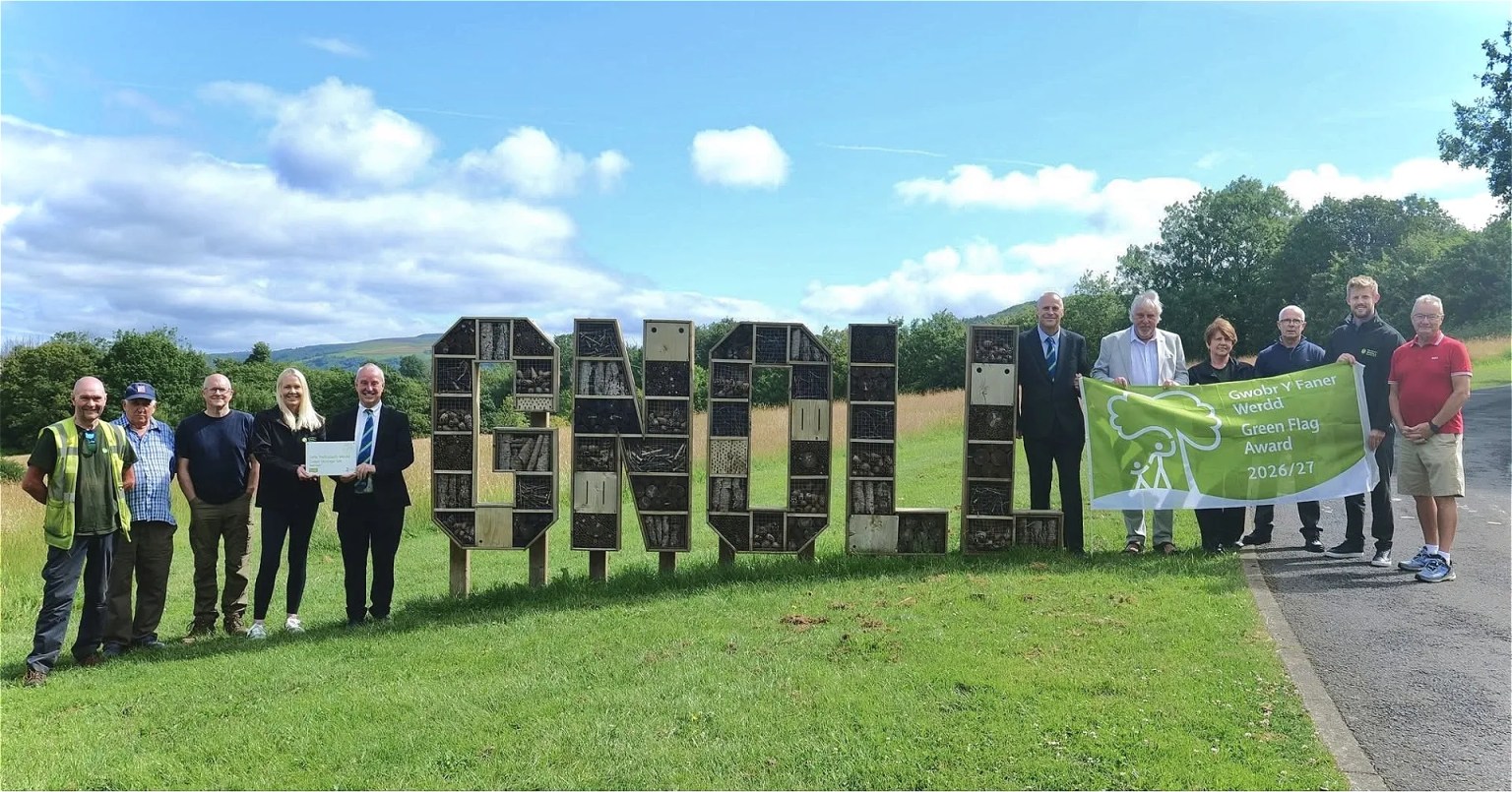 A group of people beside large wooden letters spelling GNOLL filled with bug hotels, holding a bilingual Green Flag Award 2026/27 banner.