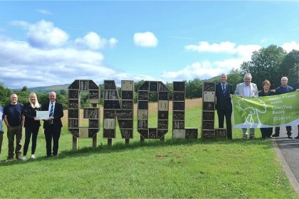 A group of people beside large wooden letters spelling GNOLL filled with bug hotels, holding a bilingual Green Flag Award 2026/27 banner.