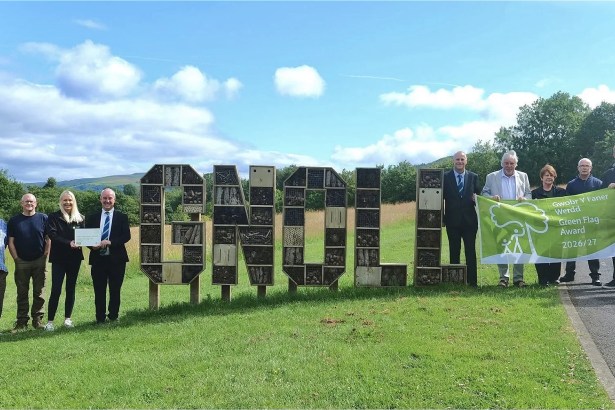 A group of people beside large wooden letters spelling GNOLL filled with bug hotels, holding a bilingual Green Flag Award 2026/27 banner.