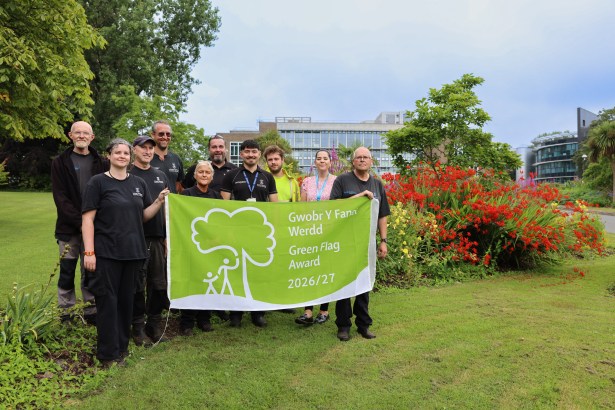 Eleven members of Swansea University's grounds team holding a green 2026/27 Green Flag Award banner in front of flower beds on Singleton campus