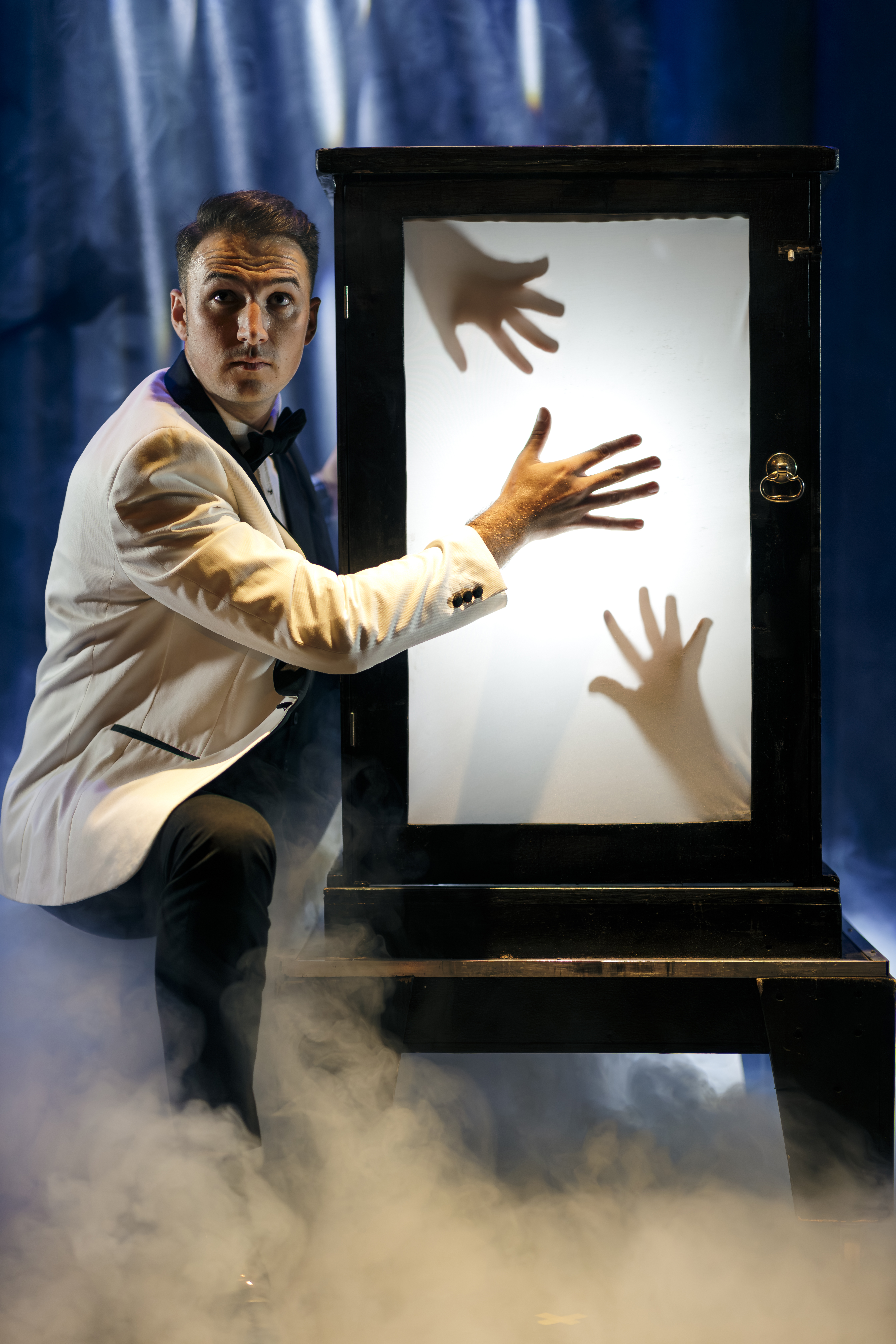 Taylor Morgan in a white dinner jacket crouching beside an illusion cabinet with shadowy hands pressing against its glowing screen