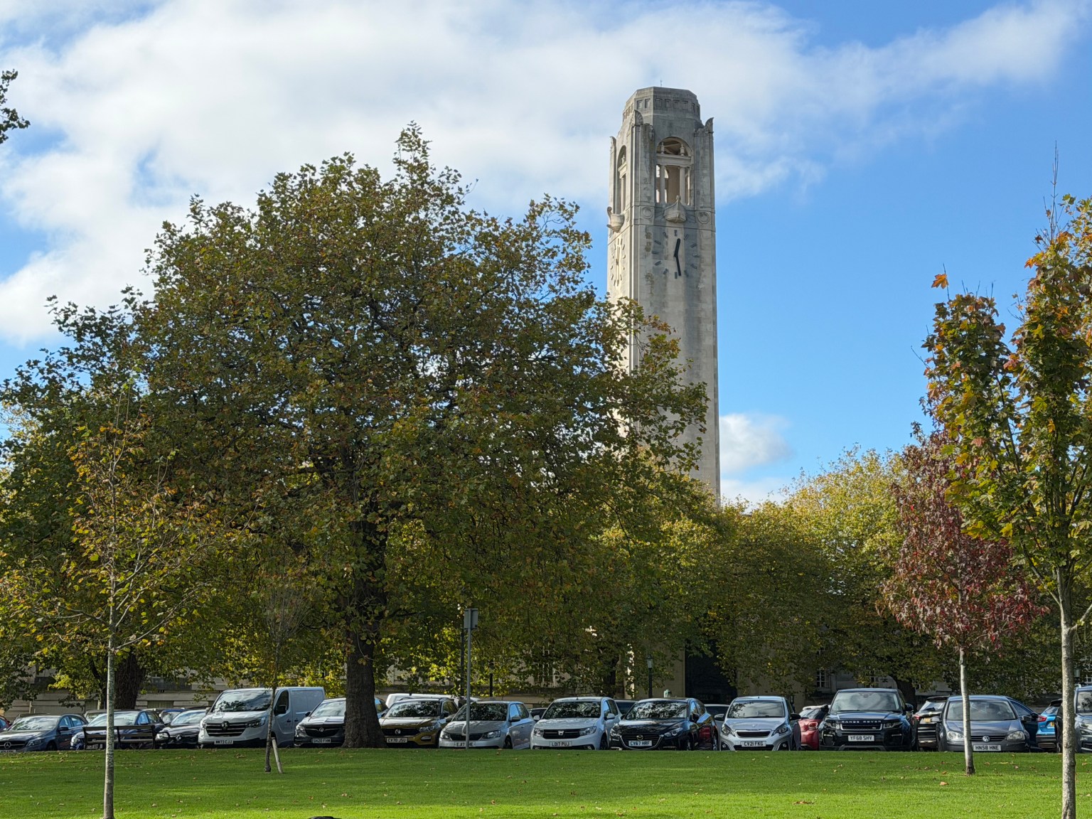 The clock tower of Swansea's Guildhall rising above trees, with cars parked in front of the building