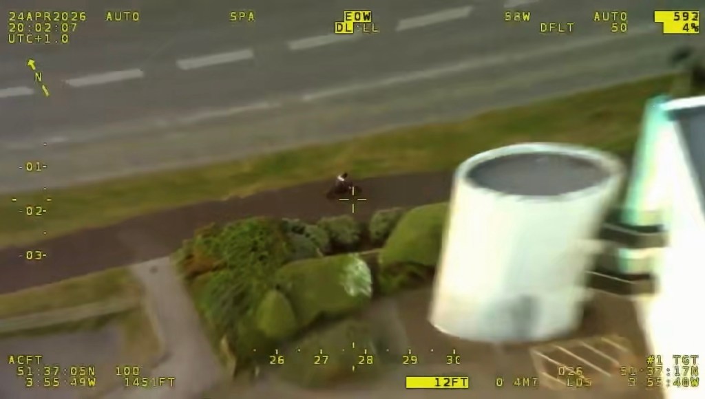 Aerial police camera view of an electric motorbike rider on a path beside a main road, near a large white storage tank