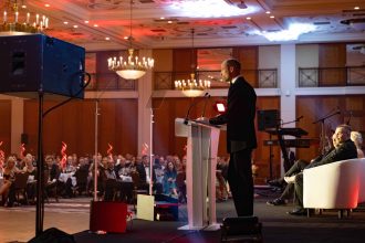 The Prince of Wales in a dinner suit speaking from a lectern on stage at a gala dinner, with guests seated at tables in a chandelier-lit ballroom.