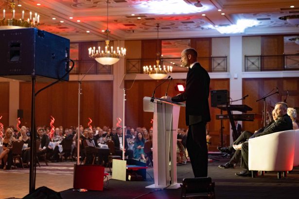 The Prince of Wales in a dinner suit speaking from a lectern on stage at a gala dinner, with guests seated at tables in a chandelier-lit ballroom.