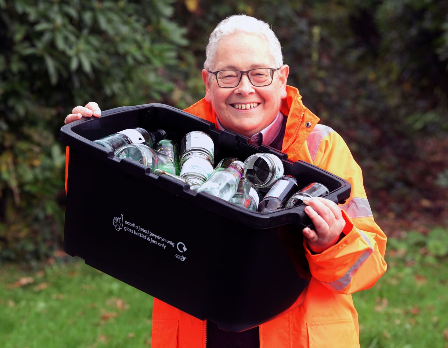 Councillor Edward Thomas in a hi-vis jacket holding a black recycling box filled with glass bottles and jars