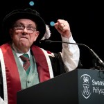 Kev Johns in academic robes raising a fist as he speaks at a Swansea University lectern