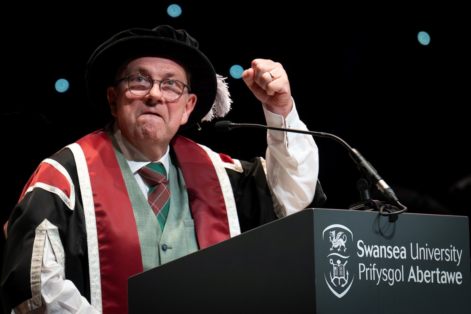 Kev Johns in academic robes raising a fist as he speaks at a Swansea University lectern