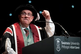 Kev Johns in academic robes raising a fist as he speaks at a Swansea University lectern