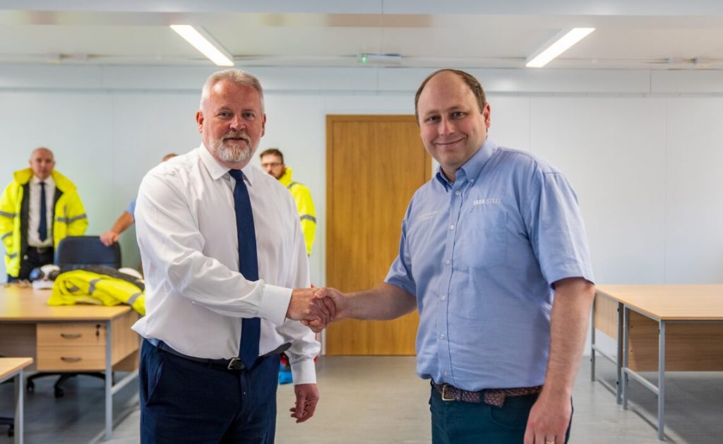Two men shaking hands in a site office, one in a white shirt and tie, the other in a blue Tata Steel shirt