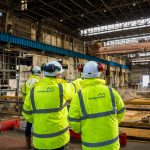 Two workers in Morganstone hi-vis jackets and white hard hats looking out across a vast steelworks building with an overhead crane and excavated foundations