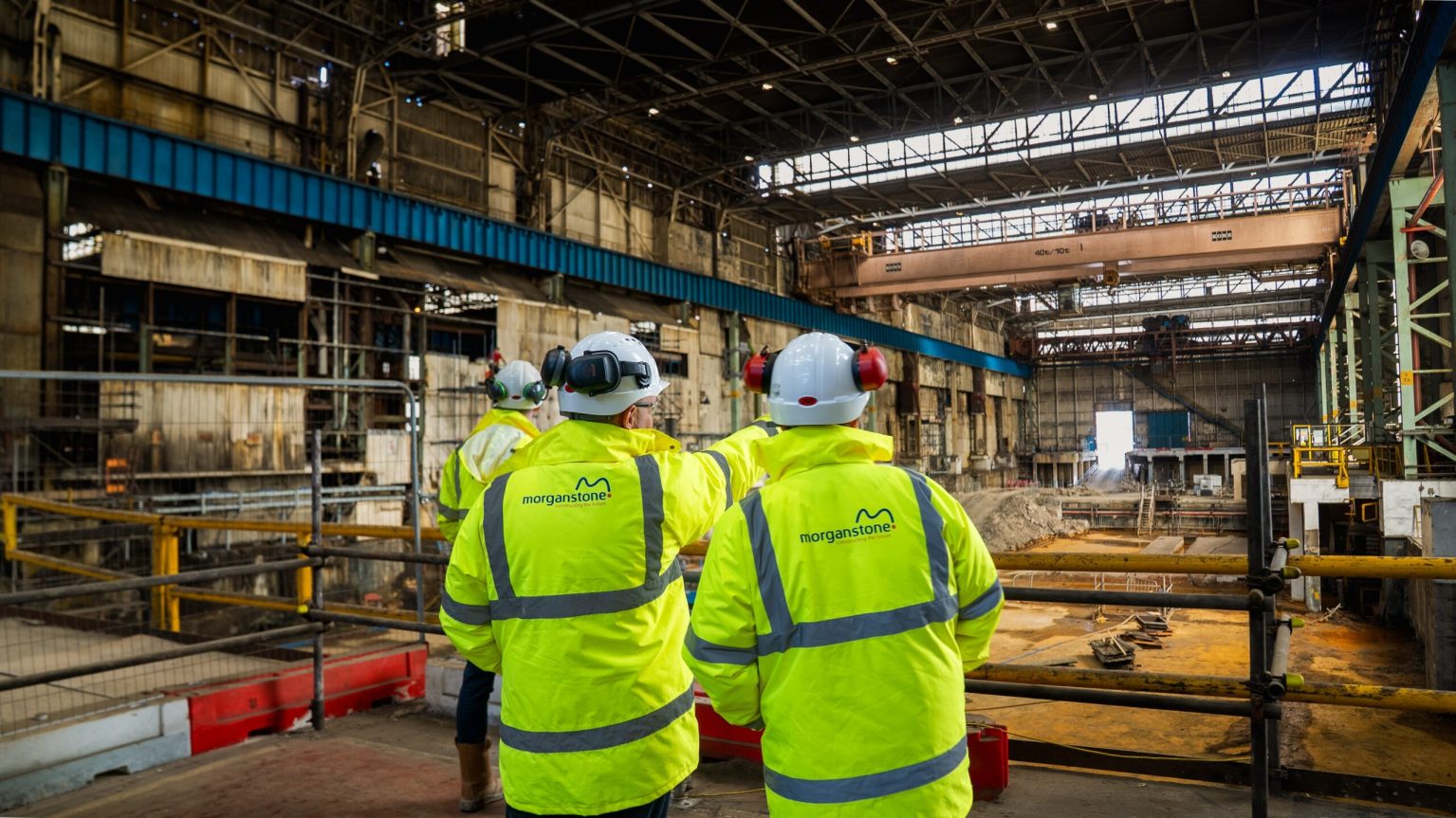 Two workers in Morganstone hi-vis jackets and white hard hats looking out across a vast steelworks building with an overhead crane and excavated foundations