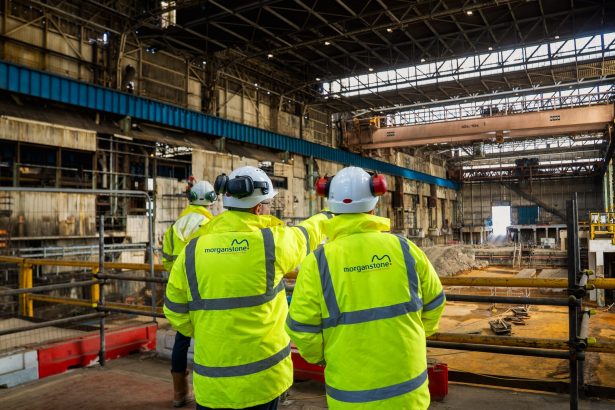 Two workers in Morganstone hi-vis jackets and white hard hats looking out across a vast steelworks building with an overhead crane and excavated foundations