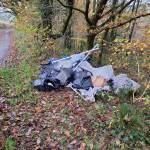 A pile of fly-tipped waste including black bin bags, plastic sheeting and metal poles on the leaf-covered verge of a country lane