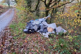 A pile of fly-tipped waste including black bin bags, plastic sheeting and metal poles on the leaf-covered verge of a country lane