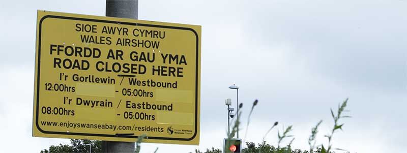 A bilingual yellow road sign reading Road Closed Here / Ffordd ar Gau Yma with airshow closure times