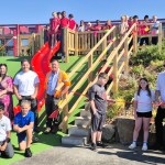 Schoolchildren and adults gathered around a red slide and wooden steps at the new SA1 park, with a pupil cutting a red ribbon and waterfront apartments behind
