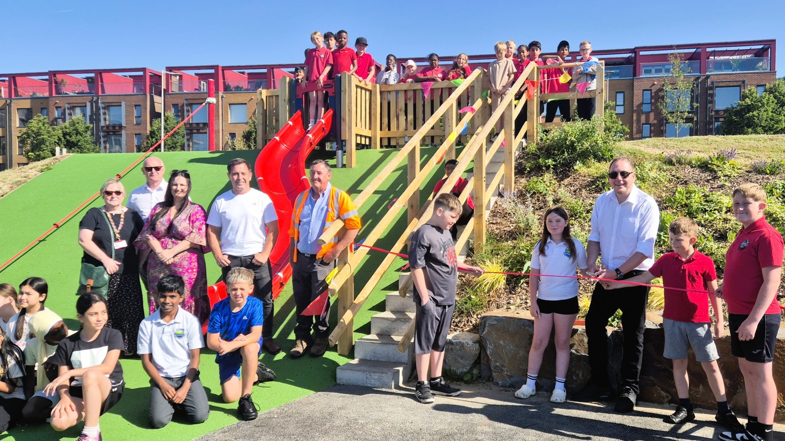 Schoolchildren and adults gathered around a red slide and wooden steps at the new SA1 park, with a pupil cutting a red ribbon and waterfront apartments behind