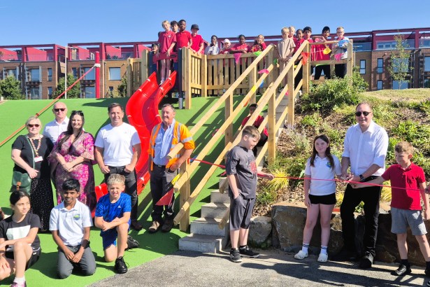 Schoolchildren and adults gathered around a red slide and wooden steps at the new SA1 park, with a pupil cutting a red ribbon and waterfront apartments behind