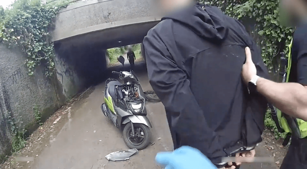A handcuffed man in a black hooded jacket held by an officer in an underpass, with a scooter and a bicycle on the path behind