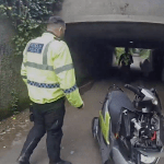 A South Wales Police officer in a hi-vis jacket with bilingual Heddlu and Police wording walks towards colleagues detaining a handcuffed man in an underpass, with a police scooter in the foreground