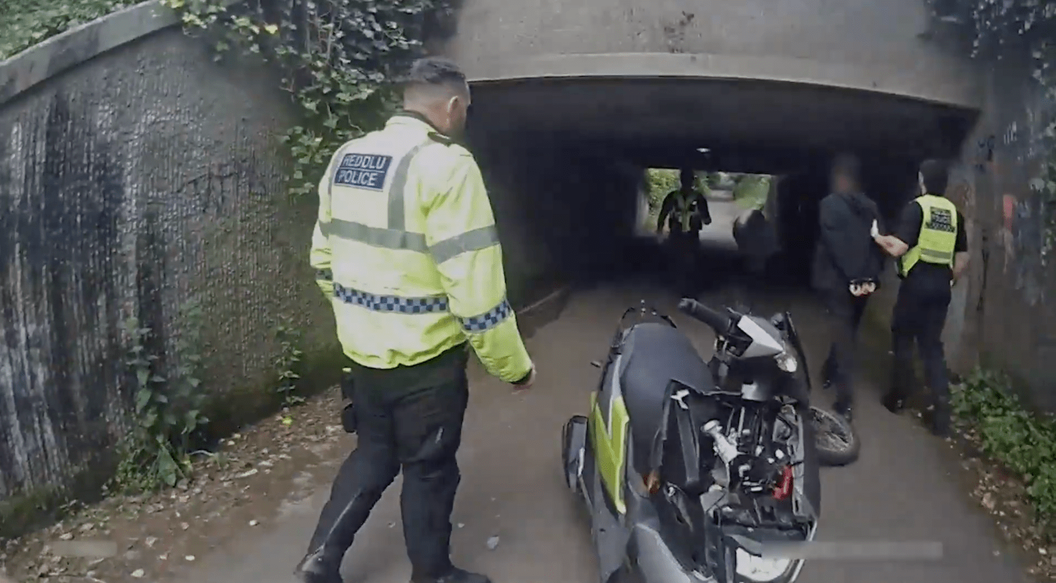 A South Wales Police officer in a hi-vis jacket with bilingual Heddlu and Police wording walks towards colleagues detaining a handcuffed man in an underpass, with a police scooter in the foreground