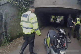 A South Wales Police officer in a hi-vis jacket with bilingual Heddlu and Police wording walks towards colleagues detaining a handcuffed man in an underpass, with a police scooter in the foreground