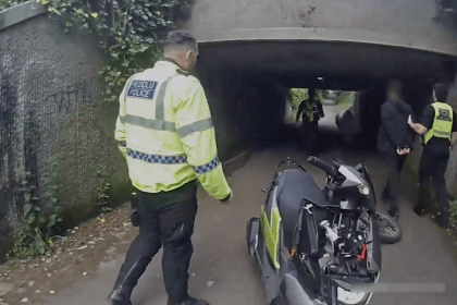 A South Wales Police officer in a hi-vis jacket with bilingual Heddlu and Police wording walks towards colleagues detaining a handcuffed man in an underpass, with a police scooter in the foreground