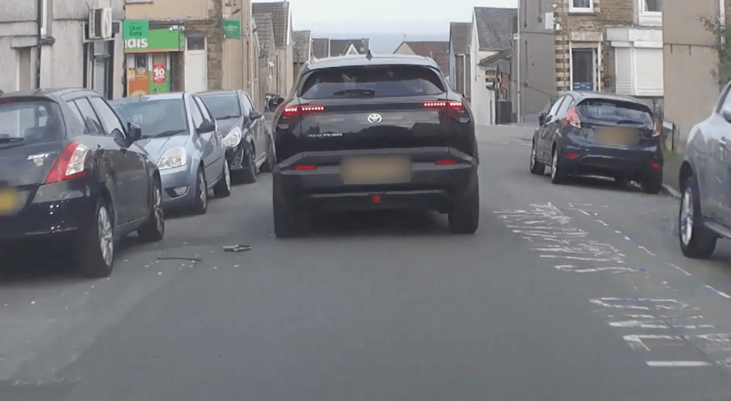 A black Toyota SUV driving between rows of parked cars on a narrow terraced street with shops at the top of the hill