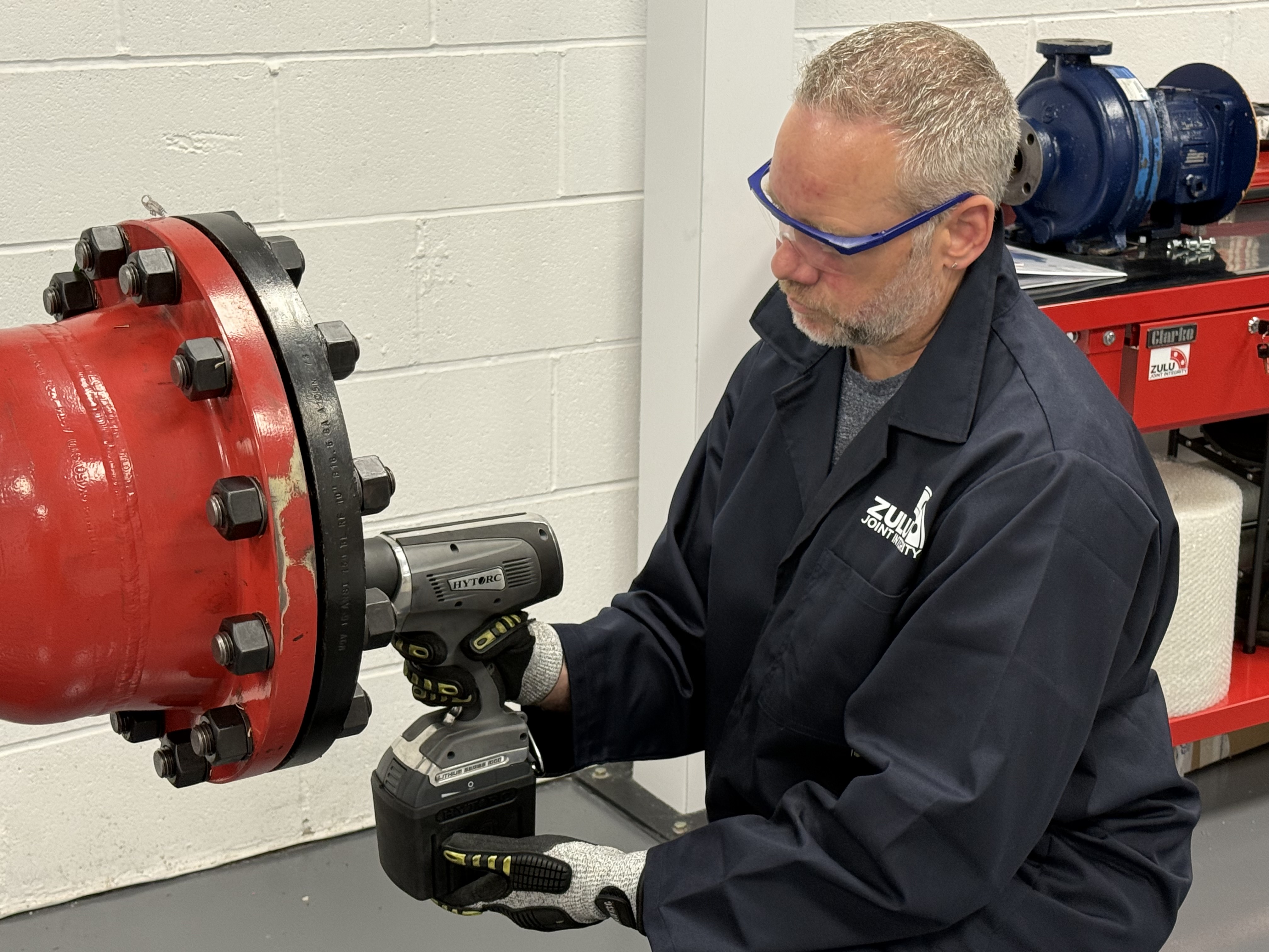 Shaun Young in navy overalls and safety glasses uses a HYTORC torque tool on the bolts of a red flanged pipe in a workshop