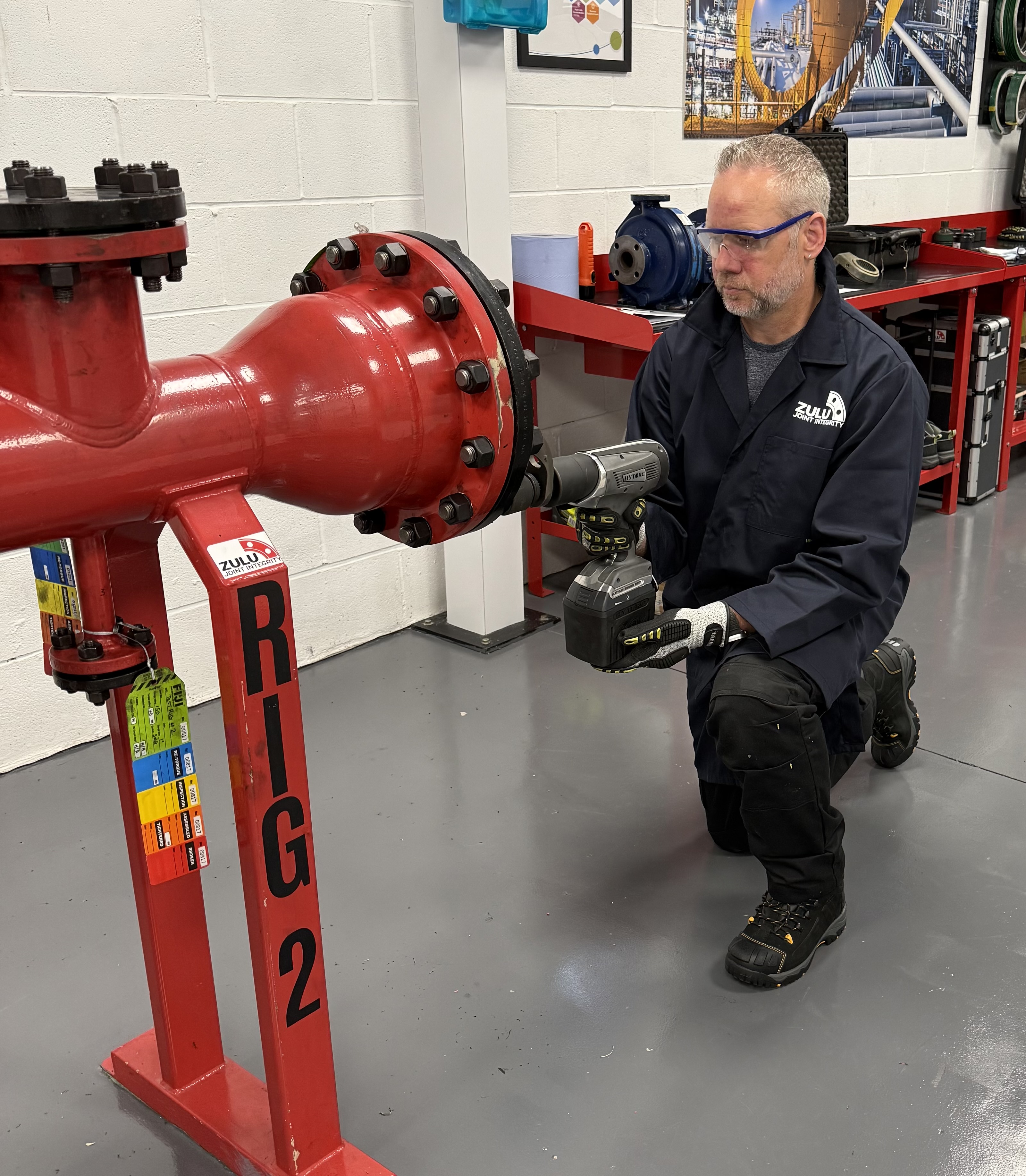 Shaun Young kneeling beside a red pipework test rig marked RIG 2, using a torque tool on a bolted flange