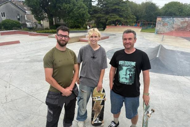 Three skateboarders holding their boards stand together at the new Victoria Park skate plaza in Swansea, with concrete ramps and bowls behind them