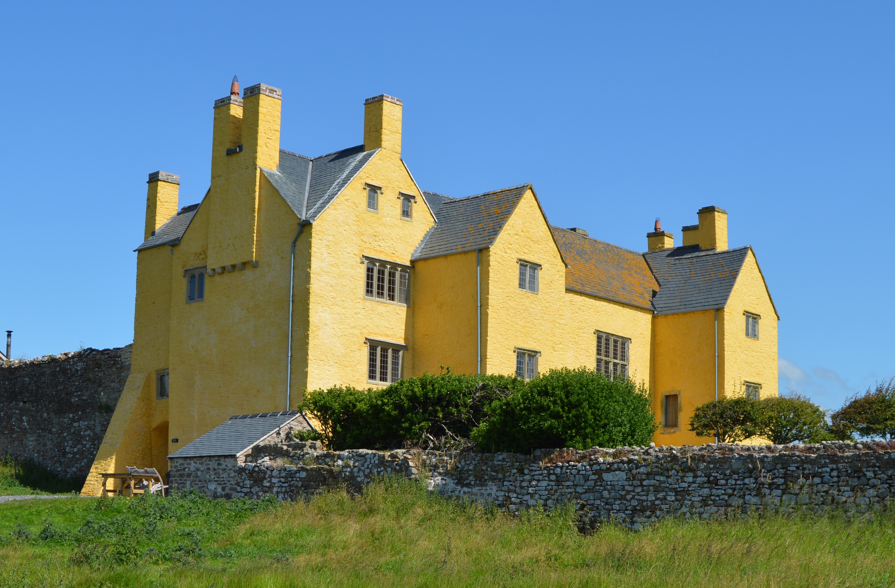 Sker House, a large yellow-limewashed Elizabethan manor with tall chimneys and slate roofs, behind a stone wall under a blue sky