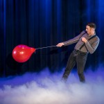 Magician Taylor Morgan on a smoke-filled stage in braces, pulling against a red balloon on a string against blue curtains