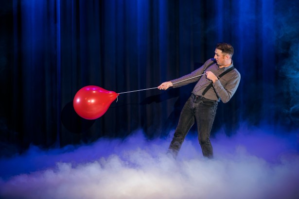 Magician Taylor Morgan on a smoke-filled stage in braces, pulling against a red balloon on a string against blue curtains