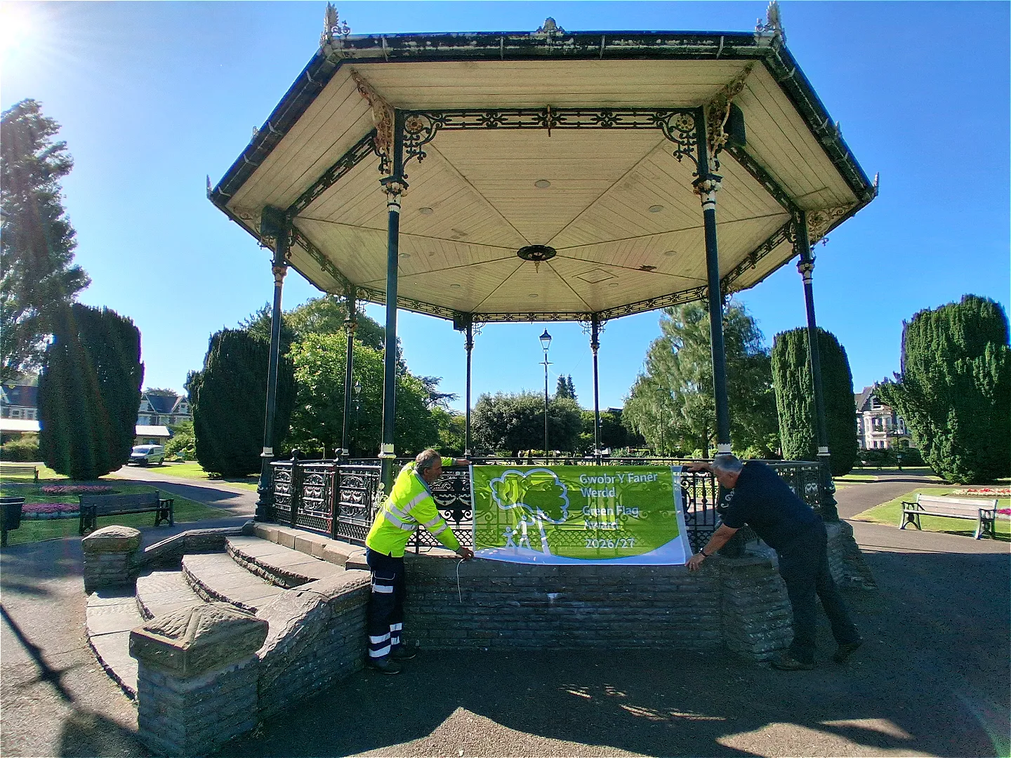 Two workers fixing a Green Flag Award banner to the Victorian bandstand at Victoria Gardens in Neath.