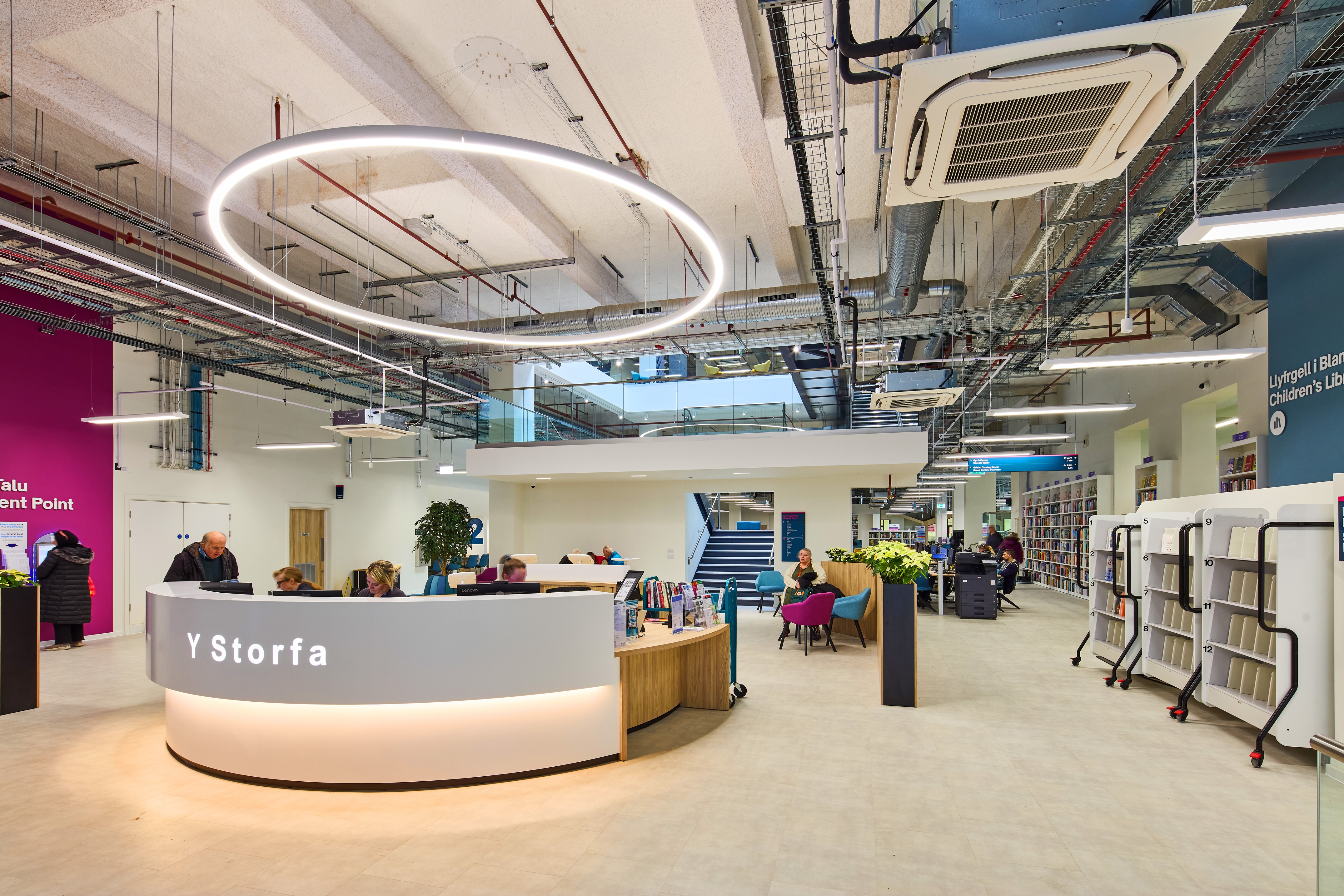 The curved Y Storfa welcome desk beneath a large circular light, with staff helping visitors and library shelves beyond