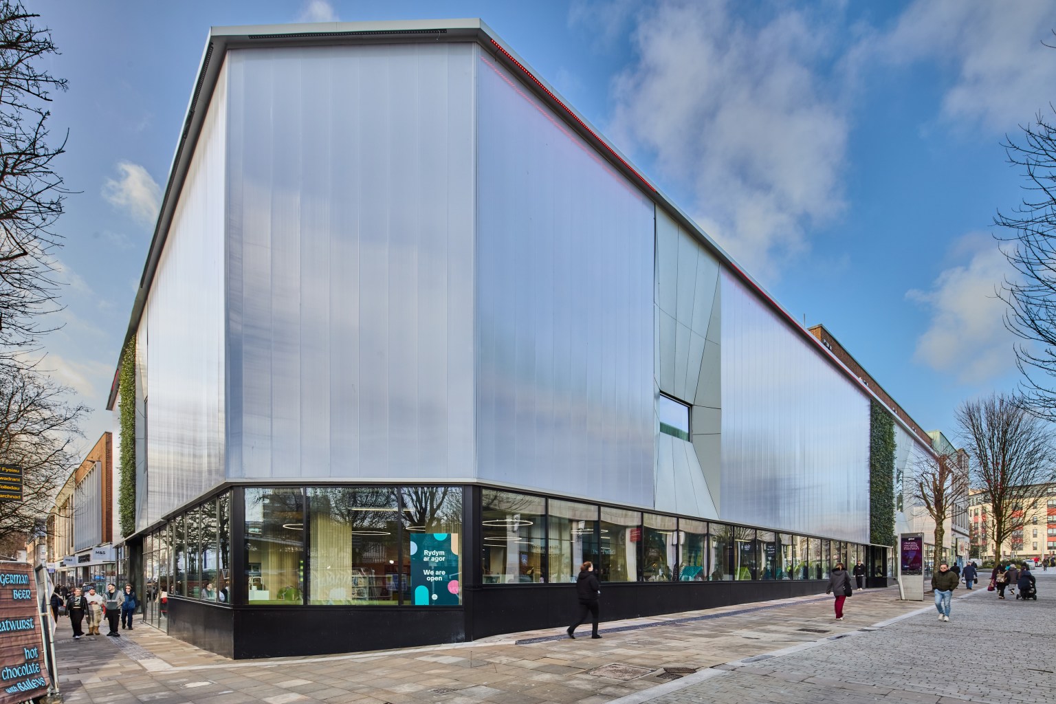 The translucent polycarbonate facade of Y Storfa in Swansea city centre, with glazed ground floor, living green walls and shoppers passing