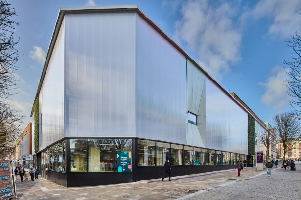 The translucent polycarbonate facade of Y Storfa in Swansea city centre, with glazed ground floor, living green walls and shoppers passing