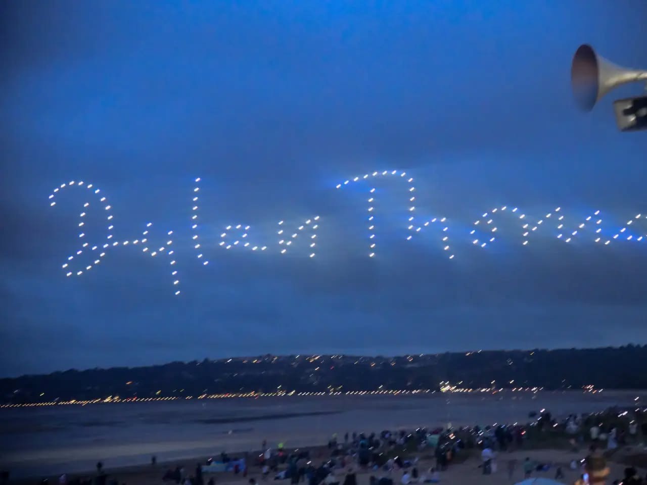 Drones spelling out Dylan Thomas in looping script above Swansea Bay, with crowds watching from the beach below
